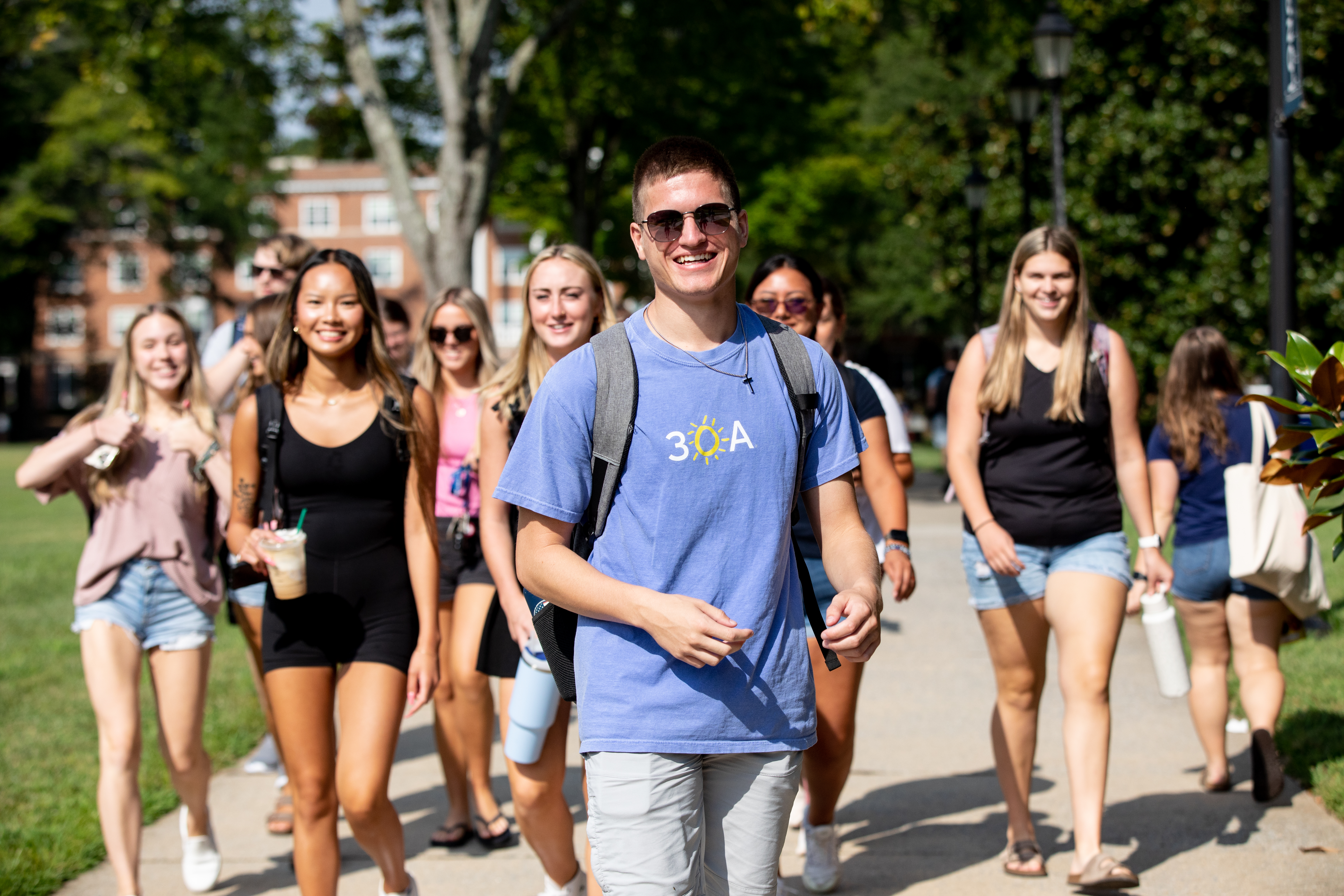 Students walk on campus in a group. The person out front is wearing a blue 3OA Beach Beach Happy shirt, aviator sunglasses and is smiling.